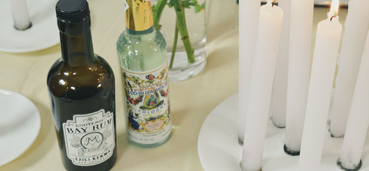 Imagen de una botella de Bay Rum rodeada de ingredientes como bayas de malagueta, jengibre, clavo dulce y canela, evocando la esencia y la preparación de esta loción caribeña.